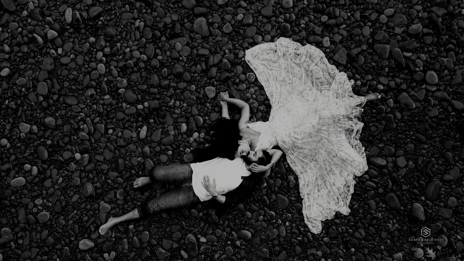 An aerial black and white frame of a couple lying on a pebble shore