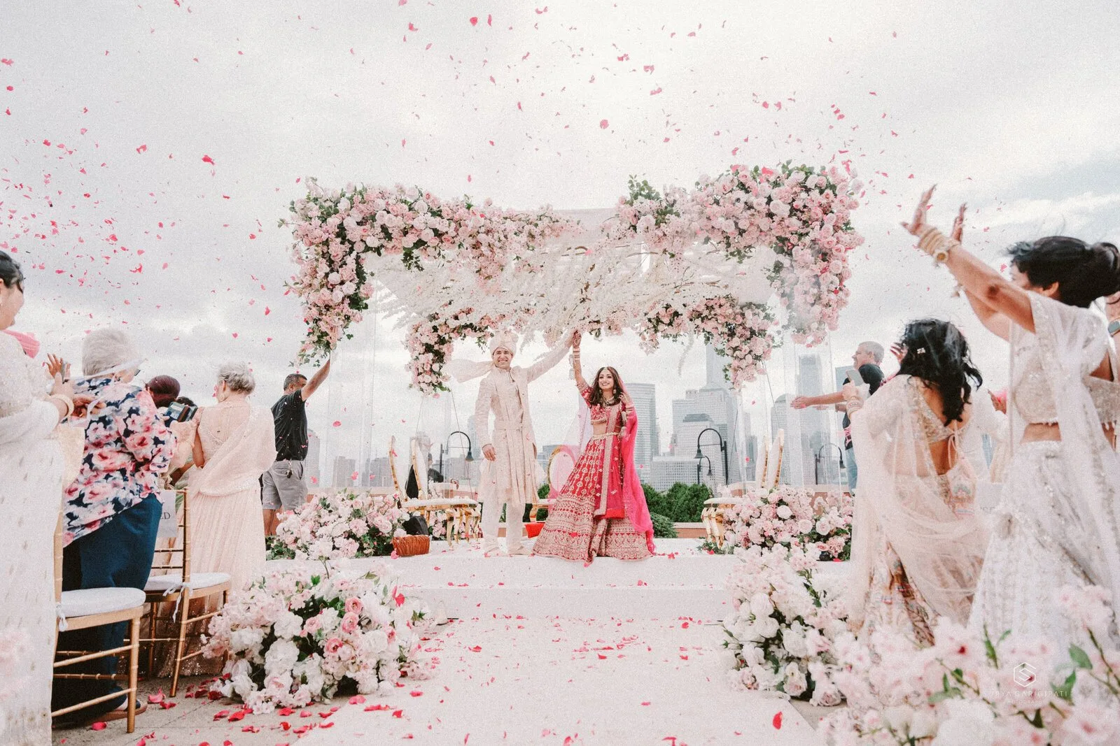 A bride and groom beneath a floral mandap with the Manhattan skyline behind them, petals in the air
